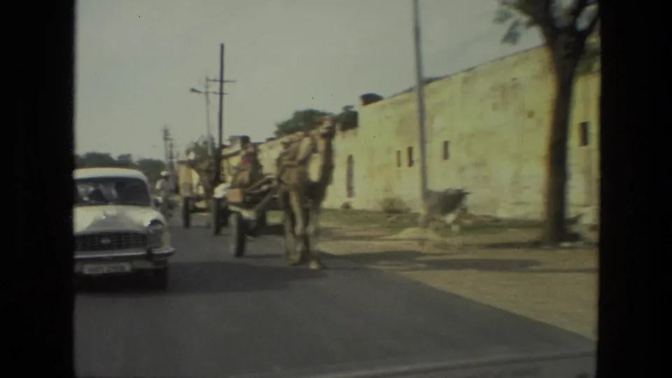 1974 India 8mm frame with a white classic car near a camel-drawn cart on a street bordered by a long masonry wall.