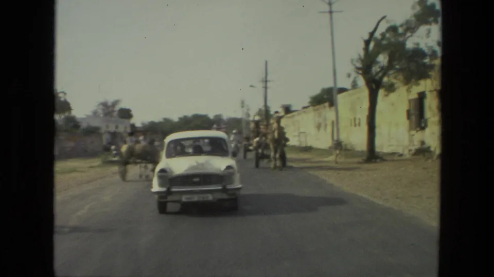 1974 India street scene with a white classic car approaching the camera, camel carts and pedestrians farther along a walled road.