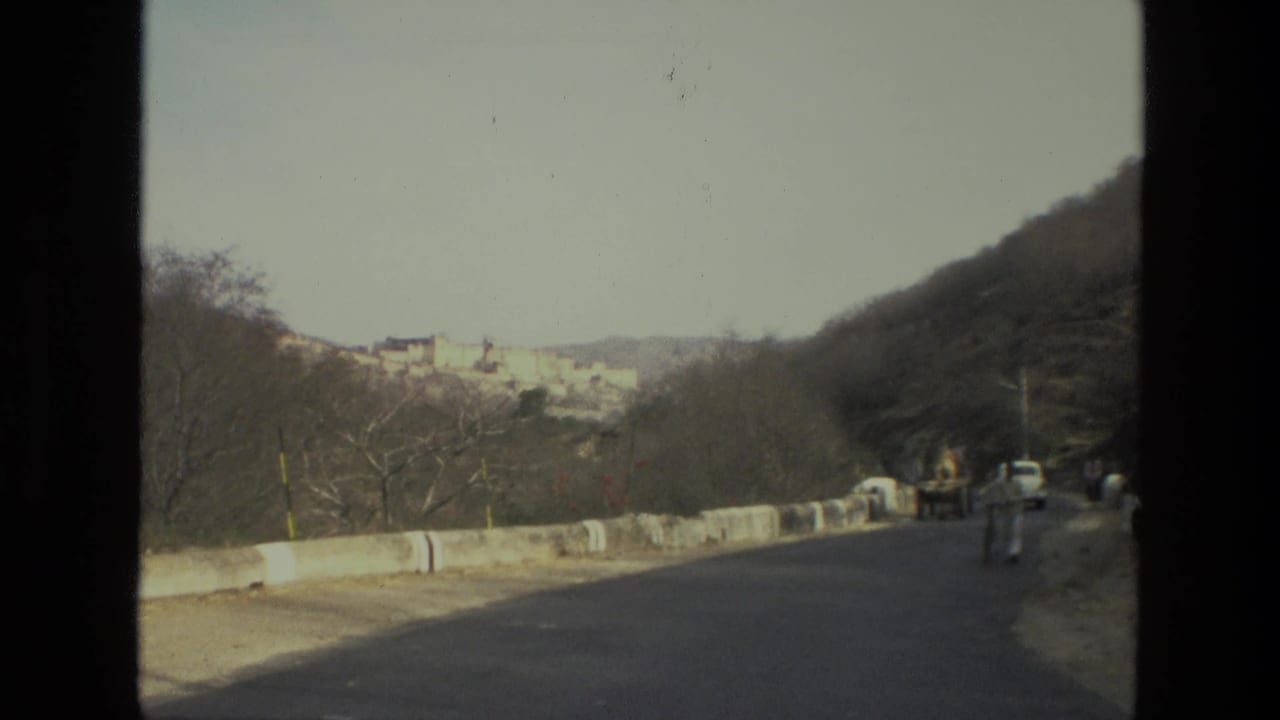 1974 India 8mm frame showing a paved rural road, roadside barrier, cyclist, white car, dry hillside, and distant palace or fort complex.