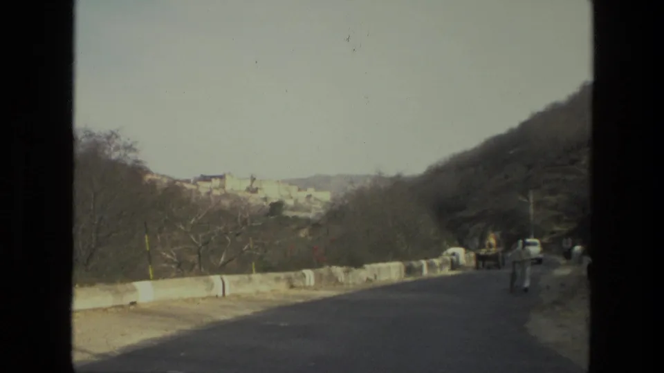 1974 India 8mm frame showing a paved rural road, roadside barrier, cyclist, white car, dry hillside, and distant palace or fort complex.