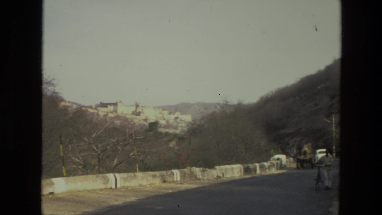 Road curves past a barrier with trees, a vehicle, and a distant hilltop complex.