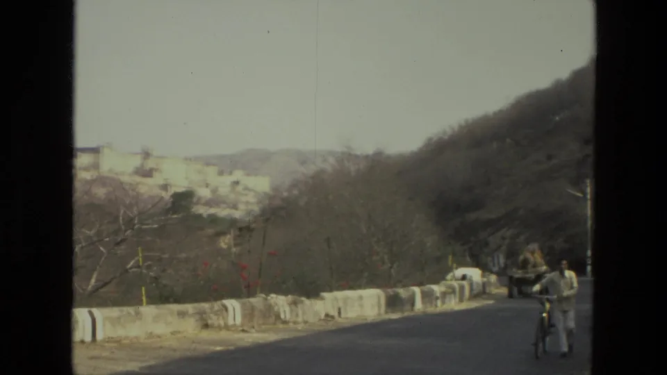 1974 India 8mm frame showing a cyclist walking a bicycle along the road, with a low barrier, bare trees, hills, and distant hilltop buildings.