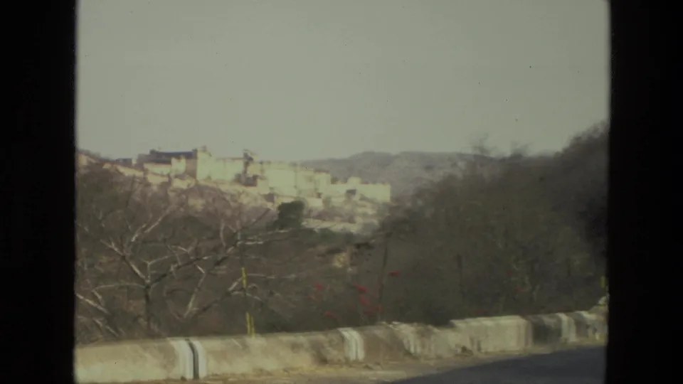 1974 India home-movie frame with a roadside barrier, leafless trees, dry hillside, and pale palace or fort complex in the distance.
