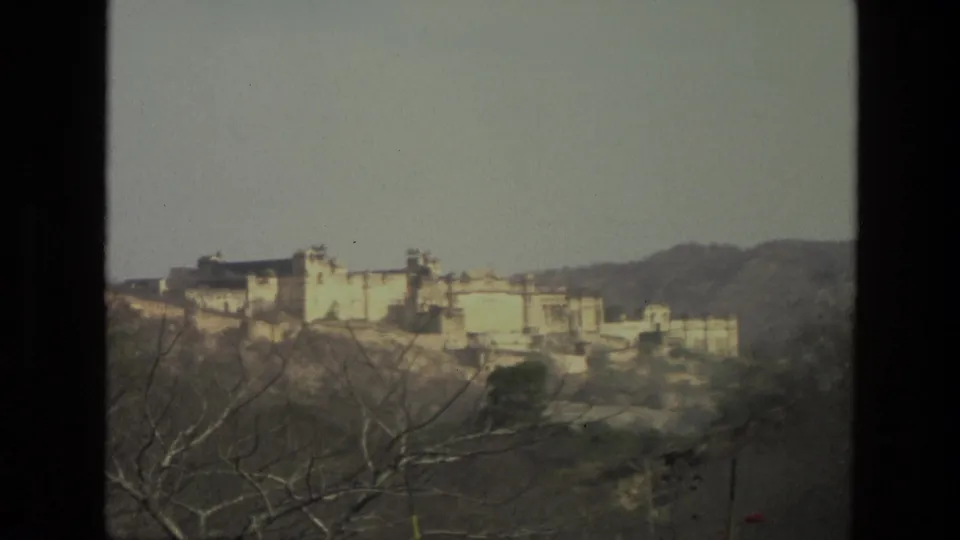 Soft 8mm frame of a distant Indian hillside fort or palace, with dry trees in the foreground and hazy hills behind.