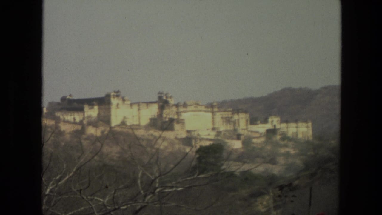 1974 India 8mm view of a pale building complex or fort-like structure on a hillside, partly obscured by branches and film edge.