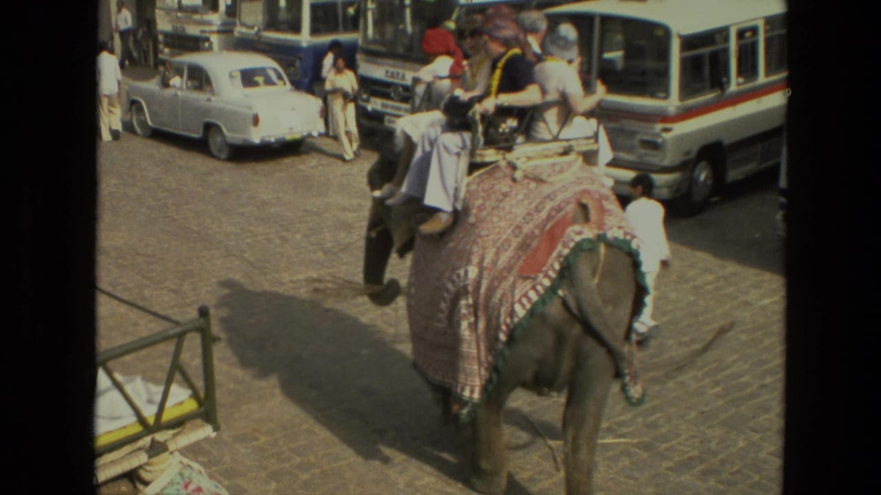 1974 India 8mm frame showing tourists riding a draped elephant through a busy street with a white sedan, bus, and pedestrians nearby.