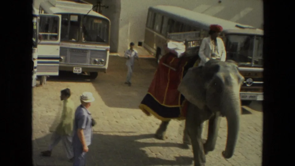 1974 India 8mm frame looking down on a decorated elephant carrying riders near buses and pedestrians on a paved street.