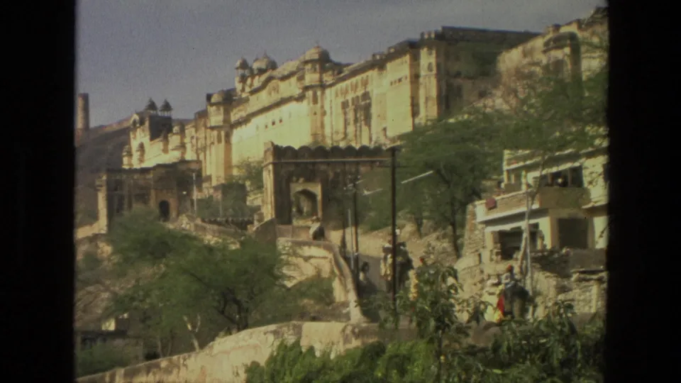 1974 India travel frame showing a large fort-like building on a hillside, with arched structures, trees, and a road below.