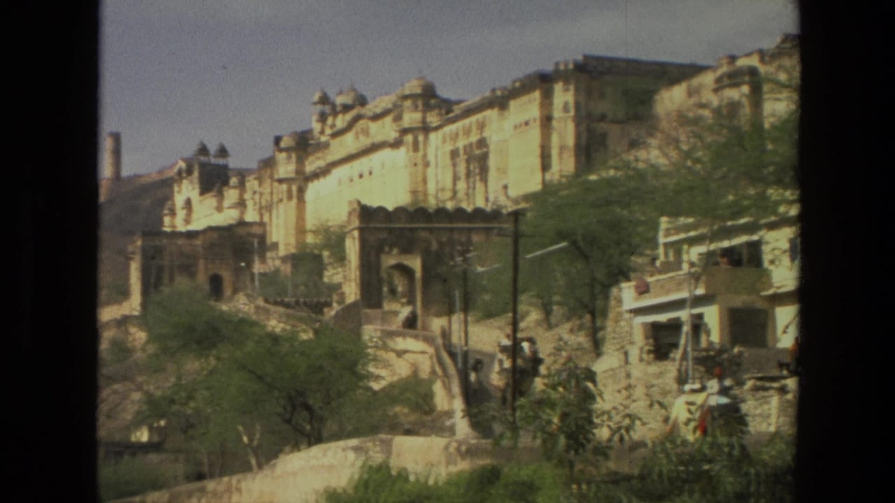1974 India frame with a large historic facade above a street, trees in the foreground, a stone wall, pedestrians, and a small roadside building.