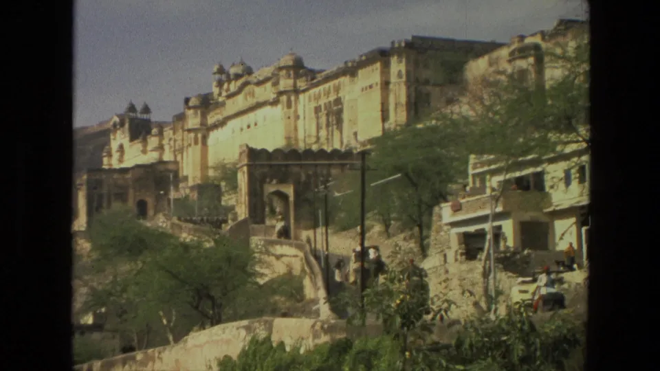 1974 India 8mm frame showing a historic hillside structure, arched gate, green trees, stone walls, and pedestrians along the street below.