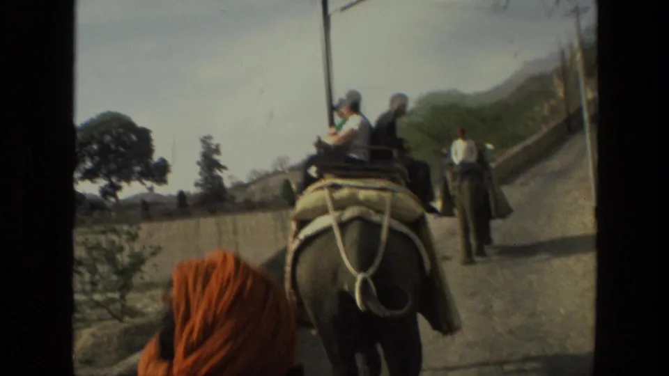 1974 India 8mm frame showing tourists on an elephant, a handler or bystander on the road, and hills beyond a wall.