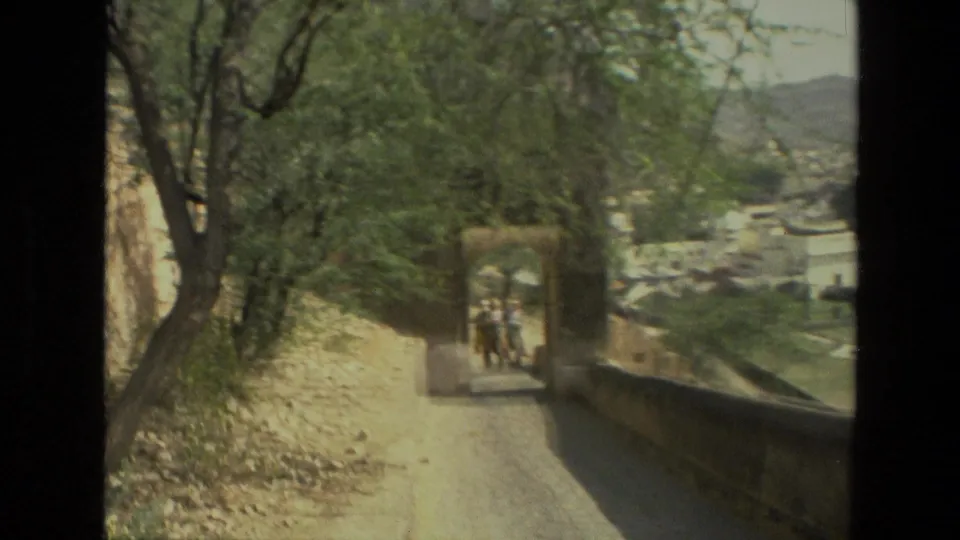 1974 India home-movie frame with an elephant and riders receding along a walled path under trees, with buildings and hills beyond.