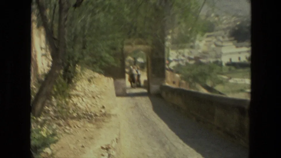 1974 8mm India travel footage showing a narrow sunlit road, trees, stone walls, and distant figures or animals ahead.