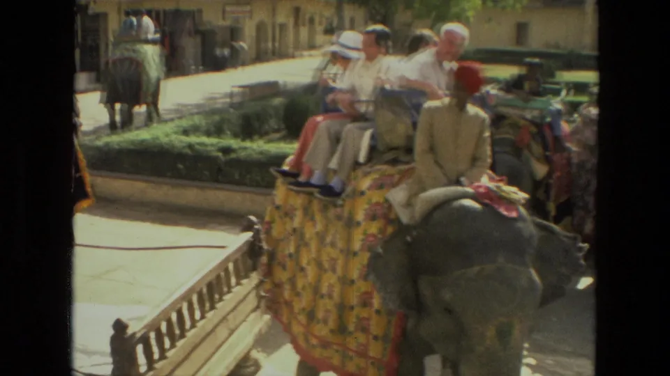 1974 India travel footage showing tourists seated on a decorated elephant beside a street railing and older buildings.