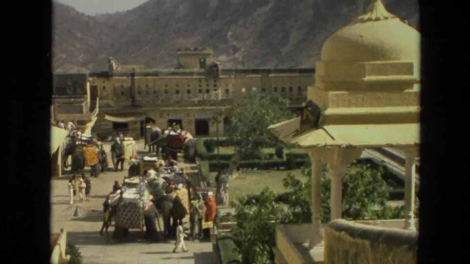 From an elevated 8mm view in India, decorated elephants and people move through a courtyard below a yellow dome where a langur is perched.