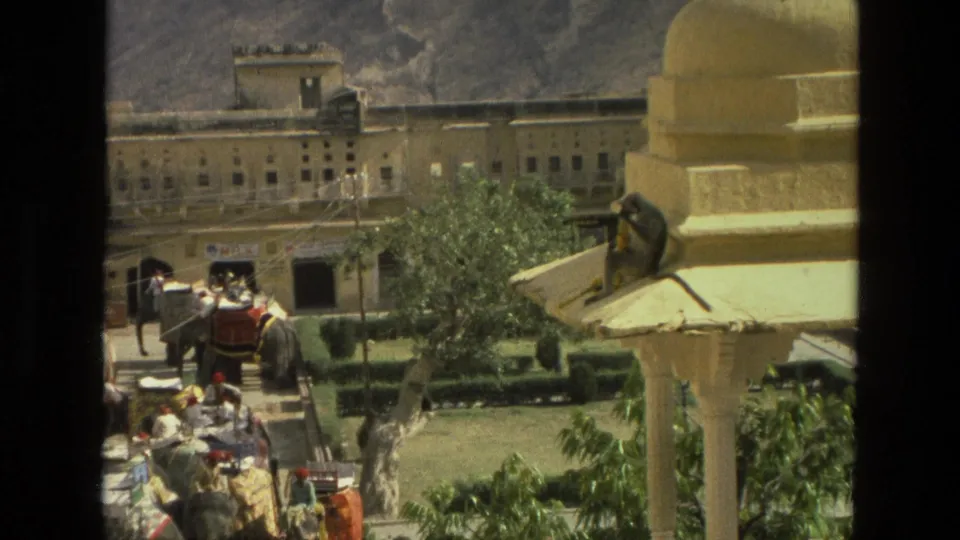 1974 India 8mm frame showing a monkey on a cream-colored roof ledge, with elephants, visitors, greenery, and a large building in the courtyard below.