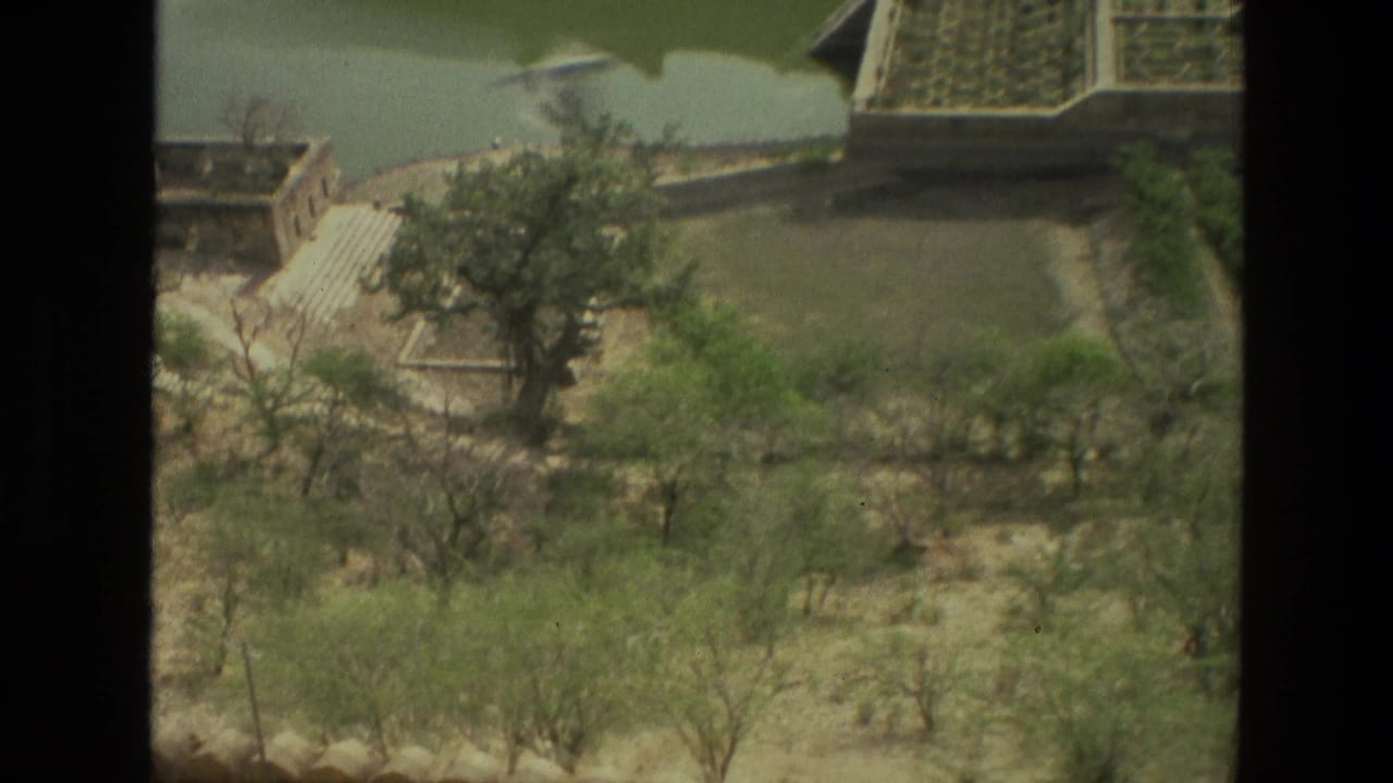 1974 8mm view in India looking down over dry landscaped grounds, scattered trees, water, and low historic-looking buildings.