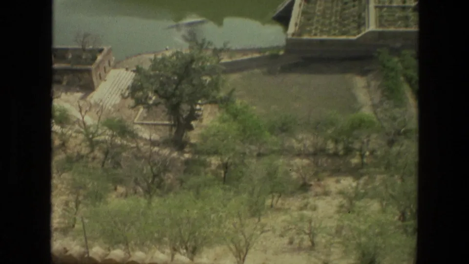 1974 8mm view in India looking down over dry landscaped grounds, scattered trees, water, and low historic-looking buildings.