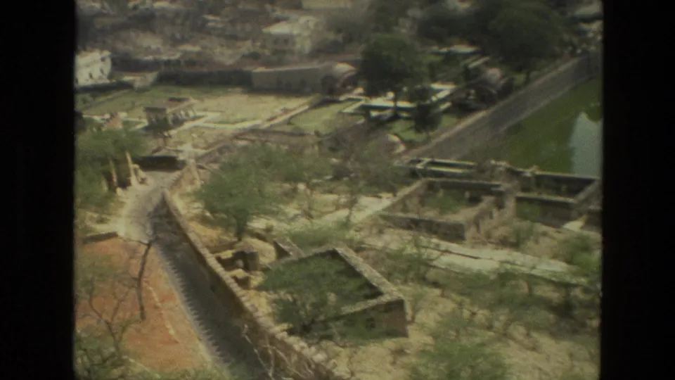 1974 India home-movie frame looking down over water, paths, dry garden plots, trees, and stone structures at a historic site.