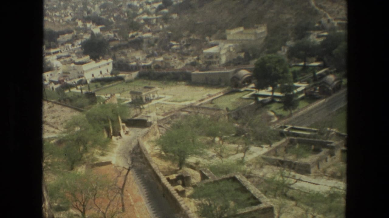 1974 8mm view in India showing a hillside city with pale buildings, fortification walls, paths, trees, and roads in a valley setting.