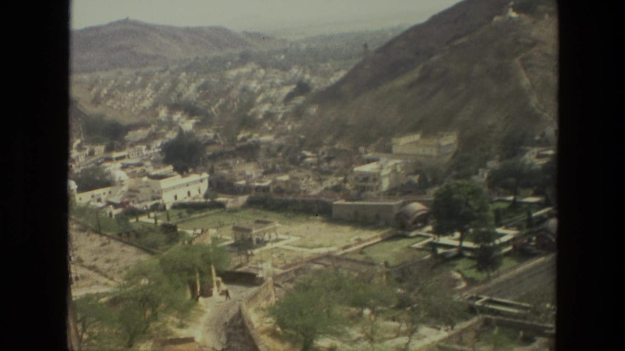 Wide valley view with clustered pale buildings, hillside roads, trees, and stone boundary walls.