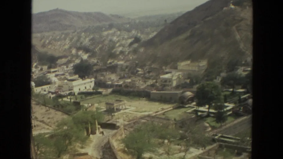 1974 India travel footage frame showing a valley city, hillside roads, pale buildings, trees, and stone walls from an elevated viewpoint.