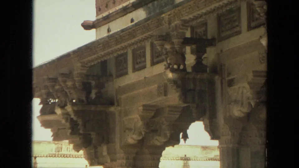 1974 India 8mm frame showing a tighter angled view of stone columns, bracket carvings, and decorative frieze work on a historic monument.