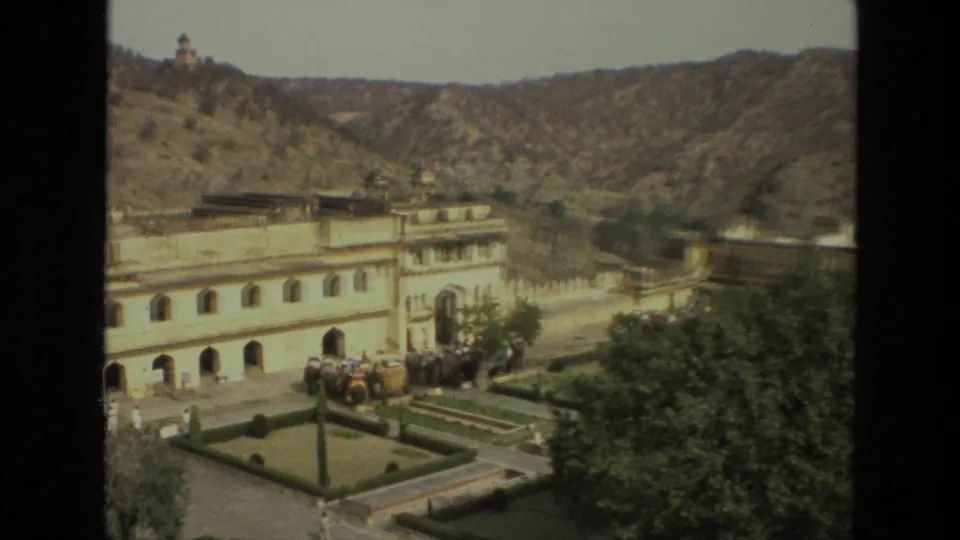 1974 India 8mm frame with a partial palace facade, landscaped courtyard, elephants, visitors, trees, and dry mountain slopes.