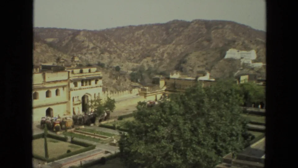 Wide 1974 India travel frame showing the palace courtyard edge, elephants, trees, rugged hills, and a distant white structure.