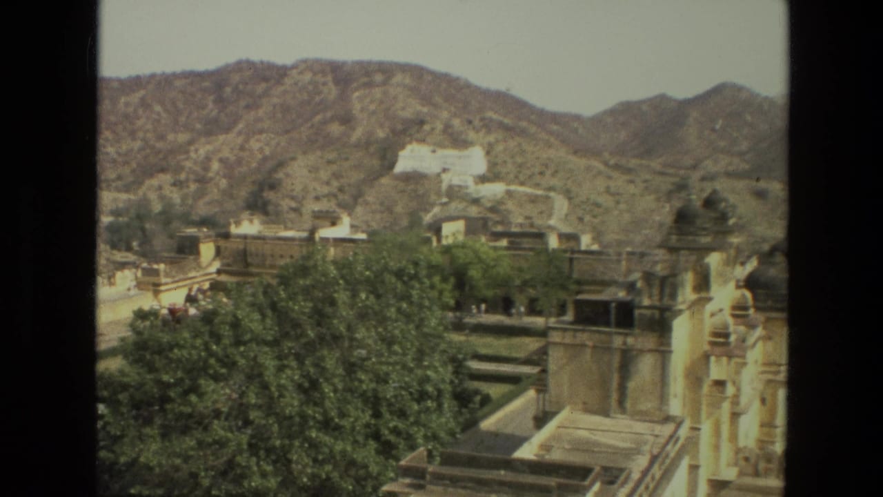 A 1974 India home-movie frame shows palace roofs, green trees, arid hills, and a distant white hilltop building.