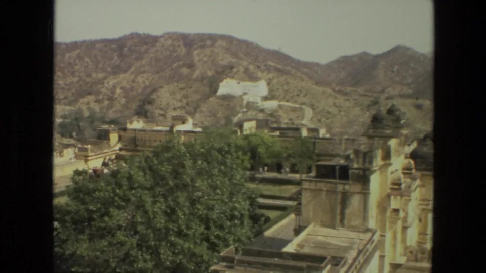 A 1974 India home-movie frame shows palace roofs, green trees, arid hills, and a distant white hilltop building.