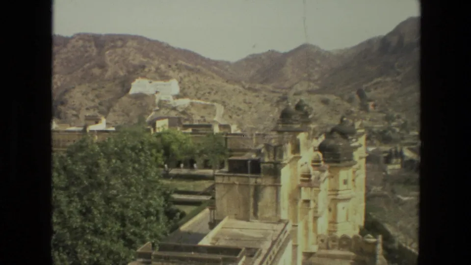 1974 India 8mm frame with domed sandstone towers, palace terraces, trees, and rocky hills in the background.