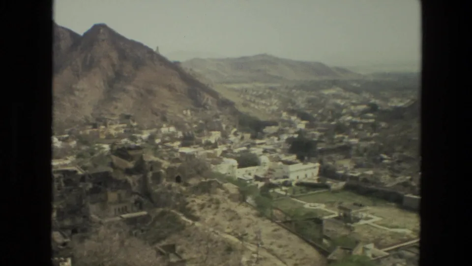 1974 India 8mm frame with town buildings, walled green grounds, and dry mountain ridges in a broad overlook.