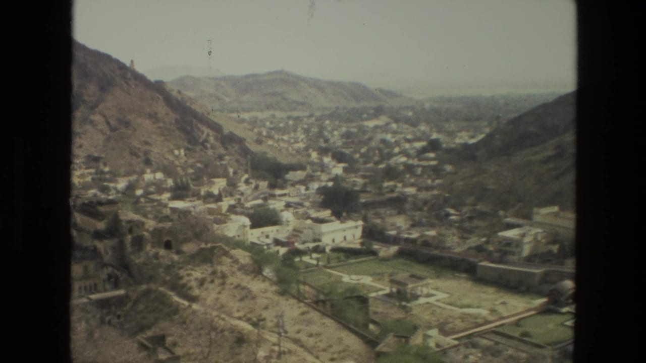 1974 8mm view in India showing a mountain valley town, clustered light buildings, roads, and walled green plots from above.