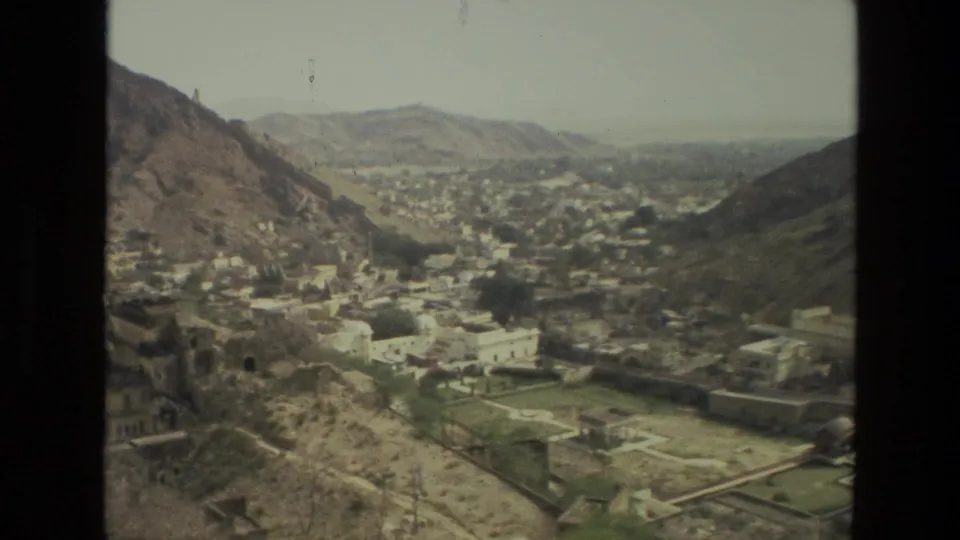 1974 8mm view in India showing a mountain valley town, clustered light buildings, roads, and walled green plots from above.