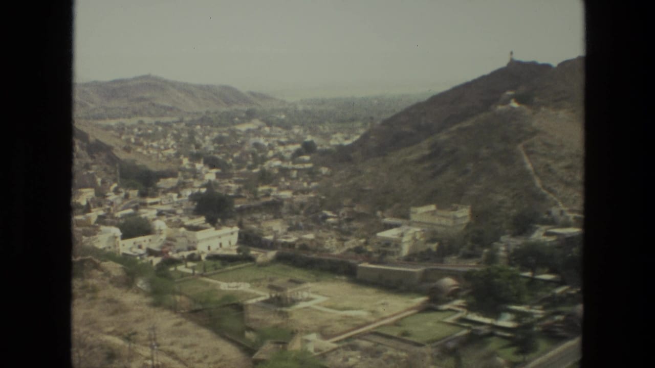 Wide valley view with clustered buildings, rocky hillsides, and a small structure high on the right ridge.