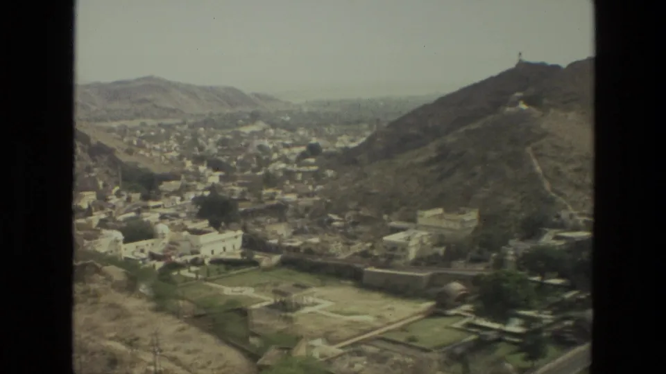 1974 India home-movie frame showing a broad valley settlement, rugged hills, and a hilltop structure on the right-hand ridge.