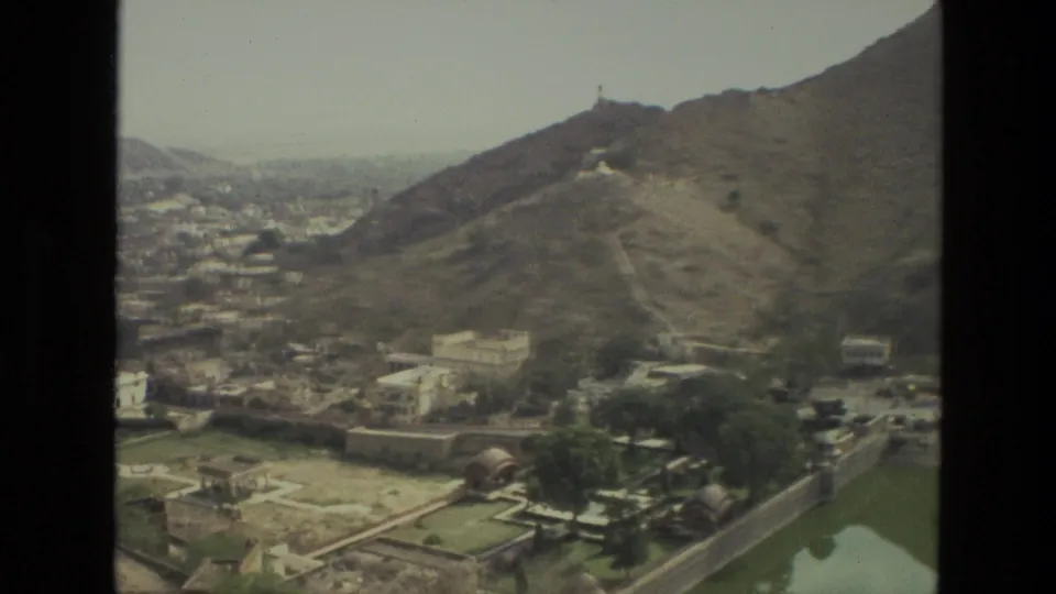 1974 8mm India frame with hillside buildings, green walled grounds, trees, water at lower right, and a small hilltop structure.
