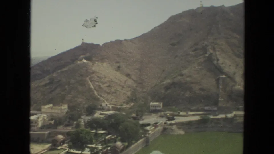 1974 India home-movie still showing a rocky hill with a winding path, small structures, trees, and green water in the foreground.