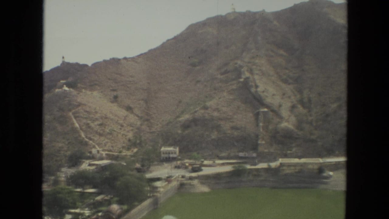 1974 8mm India travel view of arid hills, a winding path, scattered buildings, and green fields at the base of the slope.