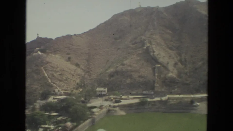 1974 8mm India travel view of arid hills, a winding path, scattered buildings, and green fields at the base of the slope.