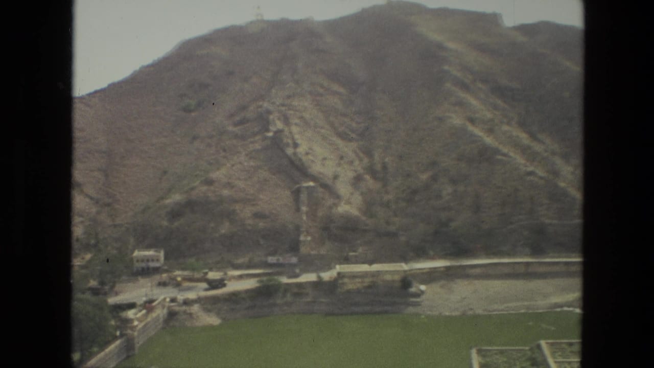 Mountain ridge with a dry channel, green field, trees, and low buildings below.