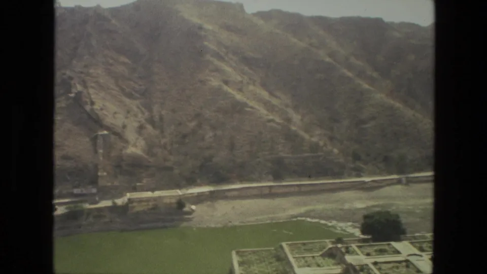 1974 8mm India view with terraced gardens, a road or embankment, green foreground area, trees, and dry hills behind.