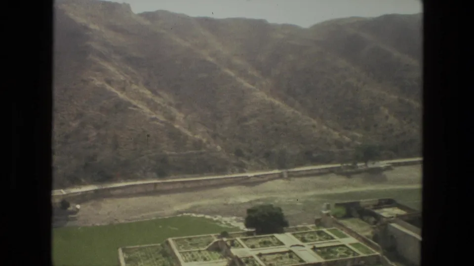 1974 8mm India travel frame showing formal garden plots, trees, a green foreground area, and a long arid hillside beyond.