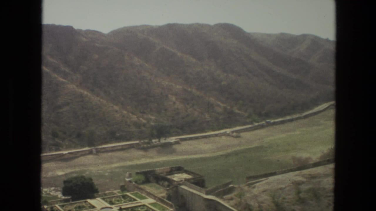 1974 India home-movie frame looking from an aircraft window across dry hills, green valley land, a road, and scattered structures.