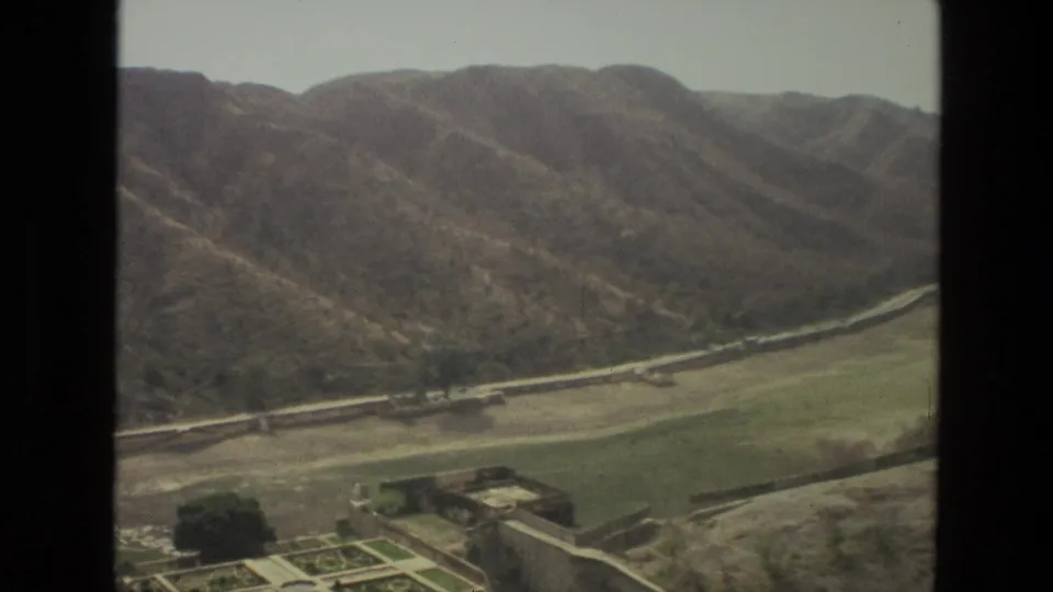1974 India home-movie frame looking from an aircraft window across dry hills, green valley land, a road, and scattered structures.