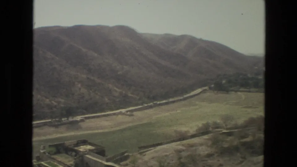 1974 8mm aerial view in India showing a winding road at the foot of dry hills, green fields, and motion-softened foreground land.