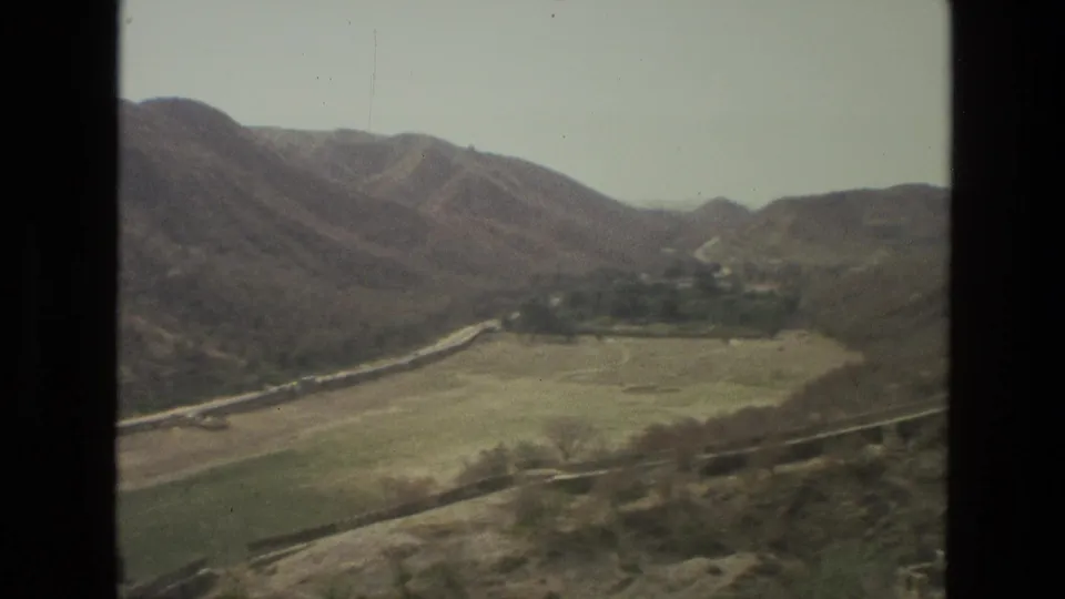 1974 India aerial home-movie frame showing a pale valley floor, a curving road, trees, and surrounding dry hills through a window edge.
