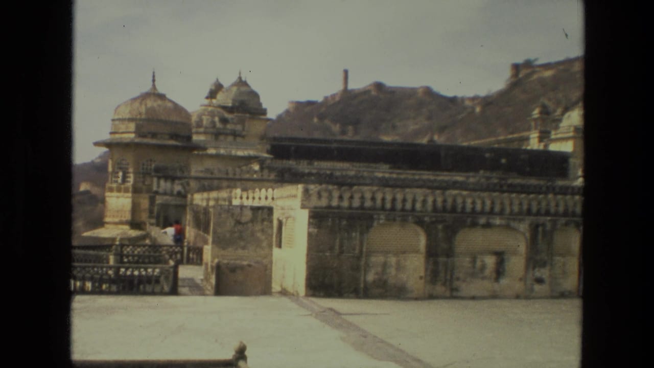 1974 India travel frame with ornate domed palace sections, a broad terrace, mountain slope, and distant visitors near the railing.