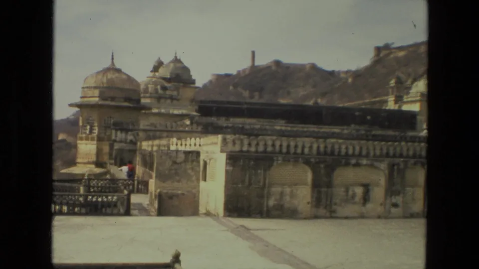 1974 India travel frame with ornate domed palace sections, a broad terrace, mountain slope, and distant visitors near the railing.